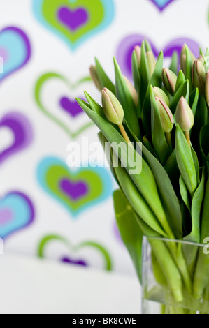 A vase of purple tulips in front of colorful wallpaper decorated with a heart pattern Stock Photo