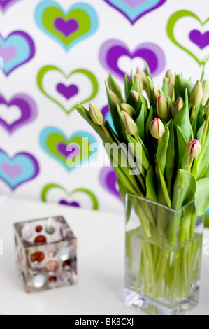 A vase of purple tulips and a glass candle holder in front of colorful wallpaper decorated with a heart pattern Stock Photo