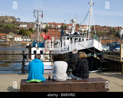 A group of sightseers one in a wheelchair sit and relax in the sunshine looking at the fishing boats at Endeavour  Wharf Whitby Stock Photo