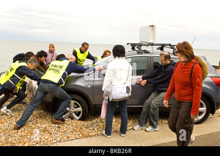 Race marshals attempt to push a car stuck in the pebbles off the beach while a woman walks past. Stock Photo