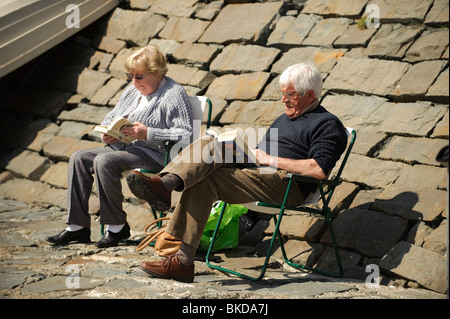 An elderly retired couple sitting in the sunshine reading books at New Quay, Ceredigion, Wales UK Stock Photo