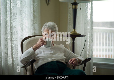 Old woman sits in living room, drinking tea from mug. Stock Photo