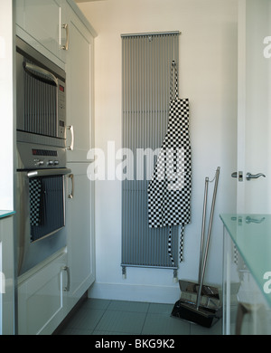 Blacked+white checked apron on vertical gray stainless steel radiator in modern white kitchen with brooms propped against wall Stock Photo