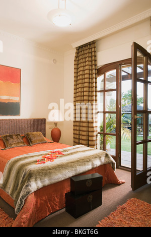 Faux-fur throw on bed with orange bedlinen in Spanish bedroom with striped curtains on open French doors to garden Stock Photo