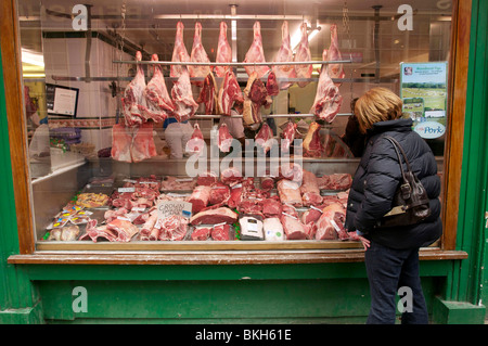 Meat hangs on display in a traditional butchers shop window with a woman looking on. Stock Photo