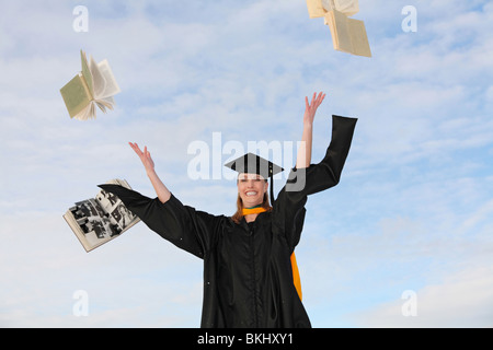 excited college graduate woman in cap and gown throwing text books into the air Stock Photo