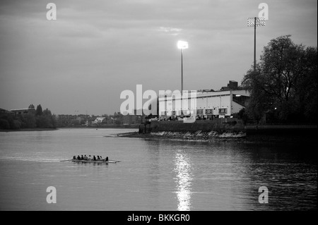 A boat full of rowers glides past Craven Cottage football ground at dusk as the floodlights glimmer on London's Thames river. Stock Photo