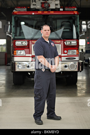 full-length fireman standing in front of fire engine at firehouse wearing blue uniform and serious face Stock Photo