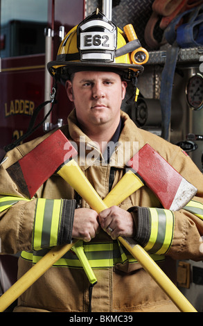 handsome middle age fireman wearing uniform and fire gear holding two axes standing in front of fire truck with serious face Stock Photo