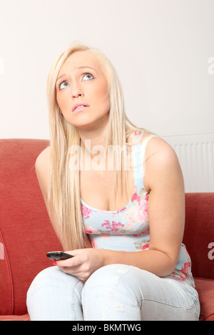 Blonde woman sitting on a red sofa looks up holding a phone. Stock Photo
