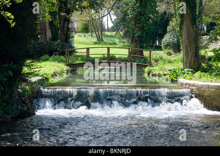 The river Glyme at Glympton, Oxfordshire, with a footbridge and small waterfall / weir, spring sunshine, Stock Photo