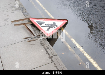 Conceptual shot of men at work sign fallen over at road side across puddle and double yellow lines. Out of work furloughed workers Stock Photo