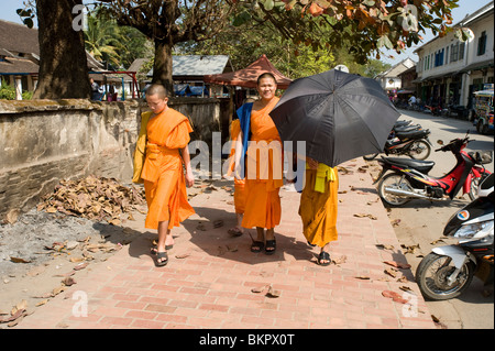 Three monks walk down the main street in Luang Prabang Laos one shielding himself from the sun with a black umbrella Stock Photo