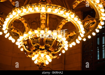 Grand Central Station Chandeliers Vanderbilt Hall New York City | NEW YORK, NY — A close view of a gilded chandelier's tiered rings of bulbs inside Grand Central Terminal's Vanderbilt Hall. Acanthus-leaf castings and gold detailing wrap the fixture's base, reflecting the Beaux-Arts craftsmanship applied throughout the terminal's 1913 construction. The angled framing reveals the hall's tall arched windows beyond. Stock Photo