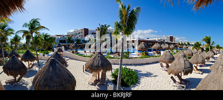 Excellence Playa Mujeres Resort Cancun Mexico | PLAYA MUJERES, Mexico — Thatched palapas and lounge chairs line the pool deck and beach at Excellence Playa Mujeres Resort, a gated all-inclusive property on the Caribbean coast north of Cancún. Playa Mujeres developed in the 2000s as a quieter, lower-density alternative to the Cancún Hotel Zone, anchored by a Greg Norman golf course and marina. The palm-fringed grounds reflect the area's contemporary resort architecture. Stock Photo