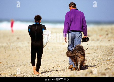 Two men walking along beach with surf board and dog Stock Photo
