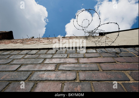 Low-angle view of a brick wall topped with barbed wire against a blue sky, suggesting security, restriction and urban tension Stock Photo