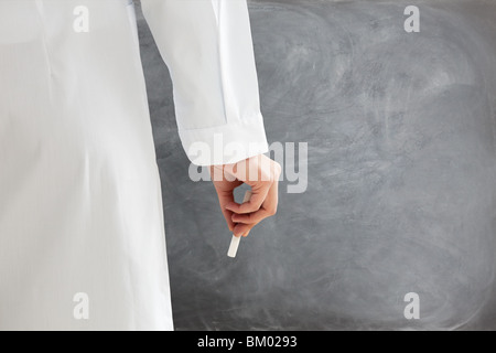 rear view of female teacher in lab clothes holding chalk against blank blackboard. Copy space Stock Photo