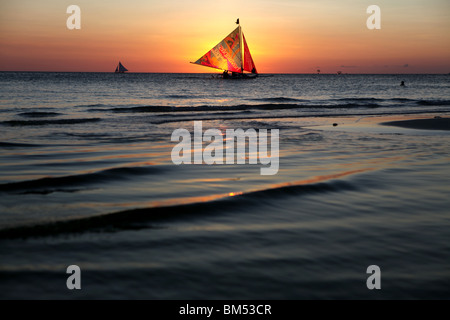 A sail boat passes the sunset on White Beach, Boracay, the most famous tourist destination in the Philippines. Stock Photo
