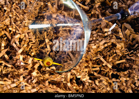 Conceptual of being empty, dry, lost. Empty martini glass setting on side in a bed of shredded brown paper with an olive @ edge. Stock Photo