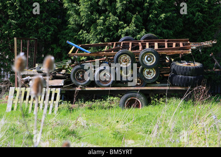 a broken trailer with rusty old trailers stacked on top, in front of conifers in the countryside on a sunny day Stock Photo