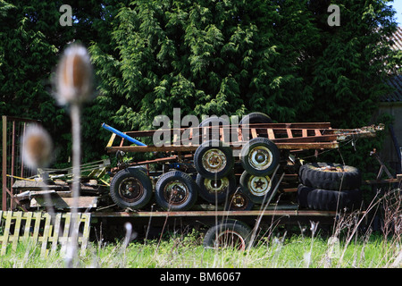 a broken trailer with rusty old trailers stacked on top, in front of conifers in the countryside on a sunny day Stock Photo