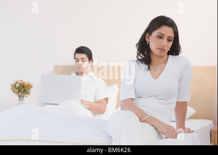 Woman thinking on a bed with her husband working on a laptop in the background Stock Photo