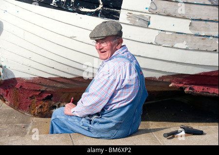 A man in his 60s in working clothes sitting in front of a fishing boat undergoing repair and maintenance Stock Photo