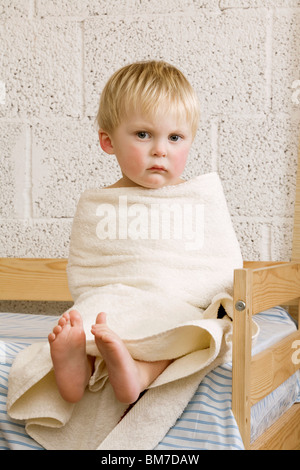 An unhappy baby boy wrapped in a towel sitting on a bed Stock Photo