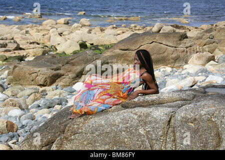 African Woman with Dreadlocks, and Wearing a Colourful Dress, Laying on Rocks by the Sea.