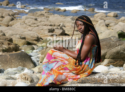 African Woman with Dreadlocks, and Wearing a Colourful Dress, Laying on Rocks by the Sea.