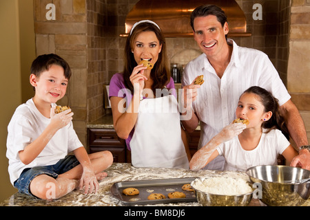 An attractive smiling family of mother, father, and two children baking and eating fresh chocolate chip cookies in a kitchen Stock Photo