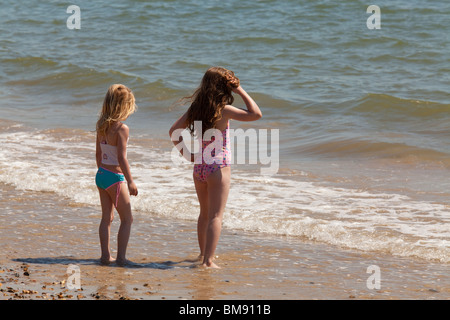 two young girls in swim suits standing together at edge of sea on beach looking out to sea