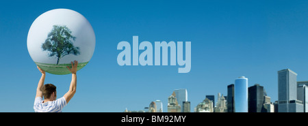 Holding aloft globe displaying image single tree, city skyline in background Stock Photo
