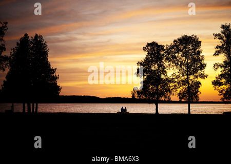 Elderly couple, silhouetted, sitting on a bench by lake at sunset Stock Photo