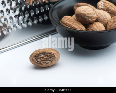 A Part Grated Nutmeg, In Front Of A Bowl Of Nutmegs and A Grater, On A White Background Stock Photo