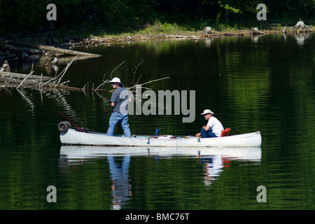 Two men in a canoe fishing along the bank of a lake one is standing. Stock Photo