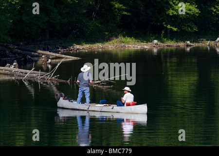 Two men in a canoe fishing along the bank of a lake one is standing. Stock Photo