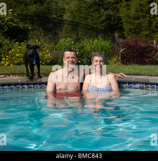 Middle aged couple, mature / retired, relaxing in a home swimming pool in the garden with a small puppy Stock Photo