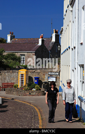 women walking on High Street of St. Anne with yelow telephone box and blue letter box, Alderney, Channel Island, United Kingdom Stock Photo