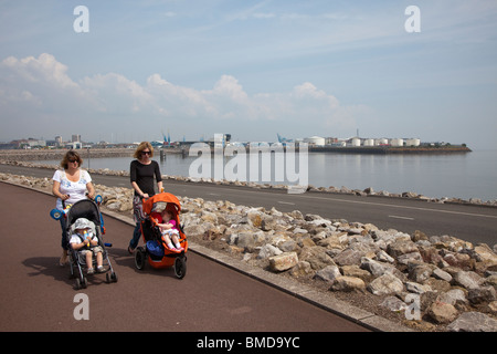 Two women pushing pushchairs walking across Cardiff Bay Barrage with oil storage tanks in docks in background Wales UK Stock Photo