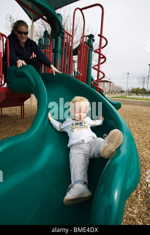 15 month old boy going down playground slide, with mother's help. Stock Photo