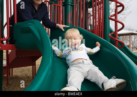 15 month old boy going down playground slide, with mother's help. Stock Photo