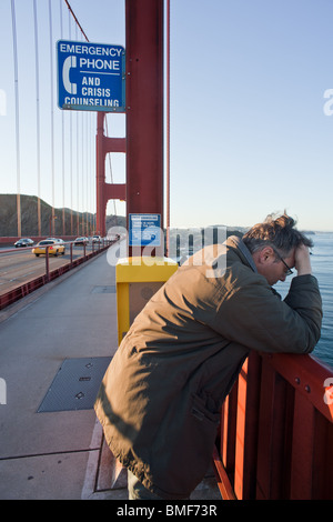 A despondent or depressed man on the Golden Gate bridge next to a suicide prevention hotline sign and phone. Stock Photo