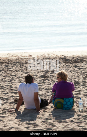 Couple sat alone on sandy beach enjoying early morning sunlight. Stock Photo