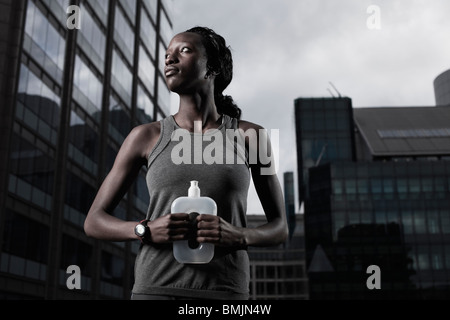 Fit young woman dressed in sportswear and holding water bottle backgrounded by modern city buildings Stock Photo