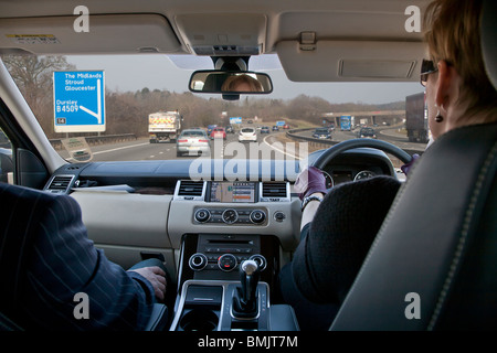 Interior of Range Rover car being driven by woman on motorway in UK, man passenger, with sat nav on dashboard and motorway sign Stock Photo