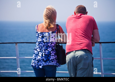 Back view of young or middle-aged couple leaning on the railing of a ferry or cruise ship looking out to sea. Stock Photo