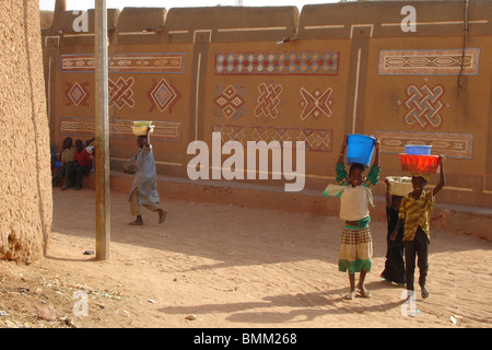 African children pausing in front of an african hausa-style house and carrying blue and red plastic buckets on the head Stock Photo