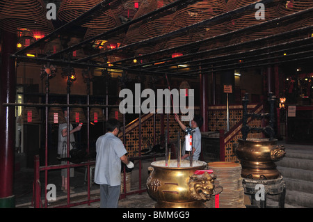 View from the side of gold incense burners and men attending incense coils hanging from the ceiling, Man Mo Temple, Hong Kong Stock Photo